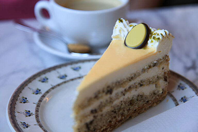 A slice of layered cake on a decorative plate with a cup of coffee in the background.