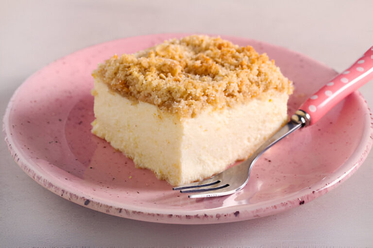 A slice of cheesecake topped with a golden crumb crust, served on a pink speckled plate with a fork.