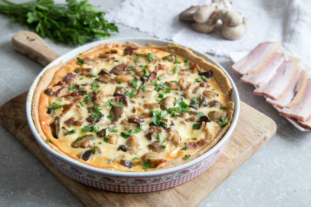 A freshly baked savory quiche with mushrooms, bacon, and parsley on a wooden cutting board, with garlic and bacon strips in the background.