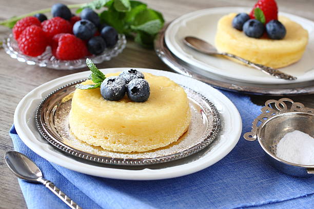 A delicious cheesecake topped with blueberries and mint on a silver plate, alongside a bowl of powdered sugar and fresh raspberries.