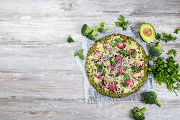 A healthy broccoli pizza topped with assorted vegetables, including avocado and herbs, on a wooden surface.