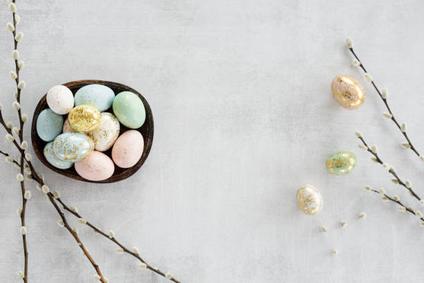 A small wooden bowl filled with pastel-colored Easter eggs on a light background, accompanied by decorative branches.