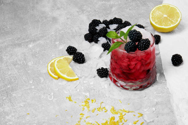 A refreshing cocktail made with blackberries, served on a textured gray surface with ice and lemon slices.