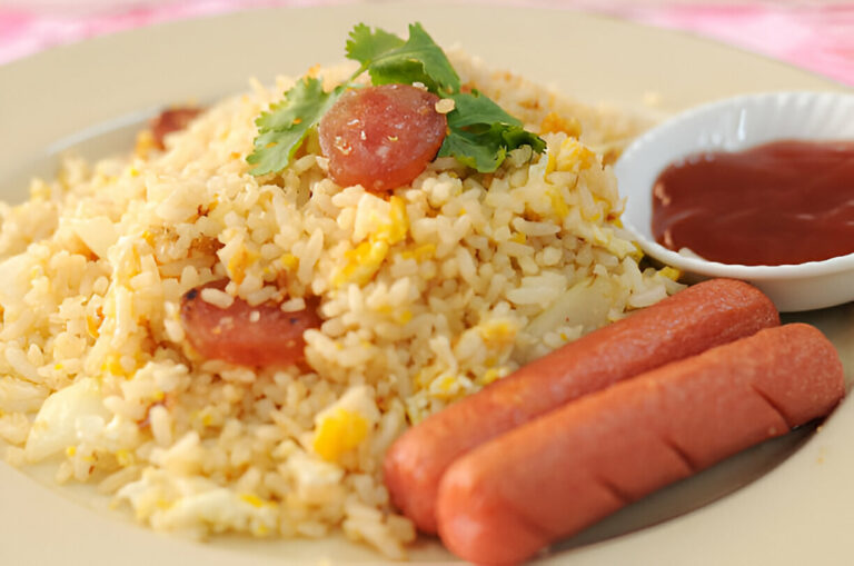 A plate of fried rice topped with sliced sausages and garnished with cilantro, accompanied by a small bowl of ketchup.