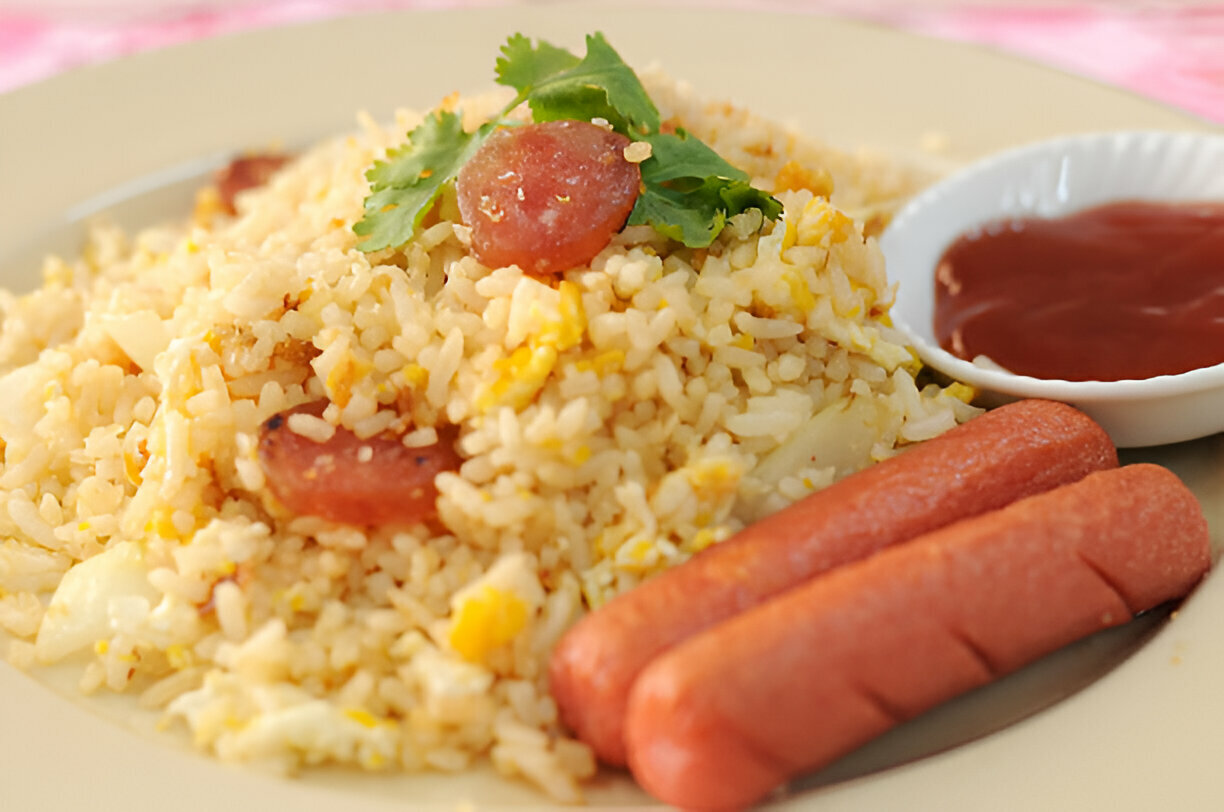 A plate of fried rice topped with sliced sausages and garnished with cilantro, accompanied by a small bowl of ketchup.
