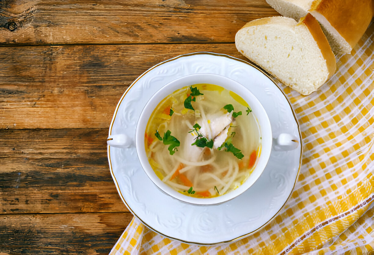 A bowl of clear chicken soup with noodles and vegetables, garnished with fresh herbs, next to a slice of bread on a checkered cloth.