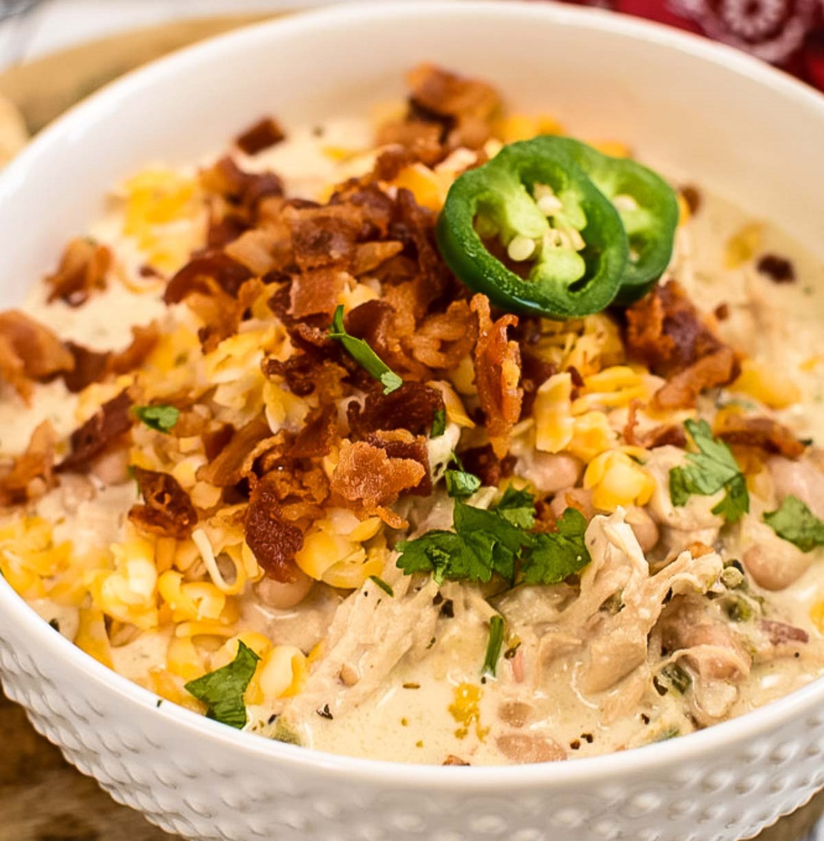 A close-up of a creamy chicken and bacon dish topped with shredded cheese, green jalapeño slices, and cilantro in a white bowl.